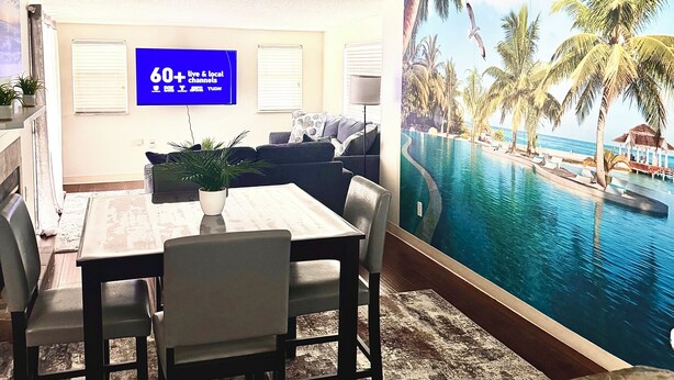 A living room with a dining area, a ceiling fan, a TV displaying '60+ live & local channels', and a large mural of a tropical pool.