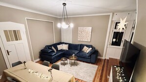 Un salon avec un canapé bleu, une table à manger avec des chaises, un lustre et un meuble pour la télévision avec des éclairages.