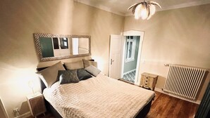 Une chambre à coucher comprenant un lit, une table de chevet, un miroir, un radiateur et une porte donnant sur un couloir.