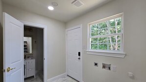 Un couloir avec une porte blanche donnant sur une salle de bain, une fenêtre offrant une vue sur les arbres et un sol en marbre.