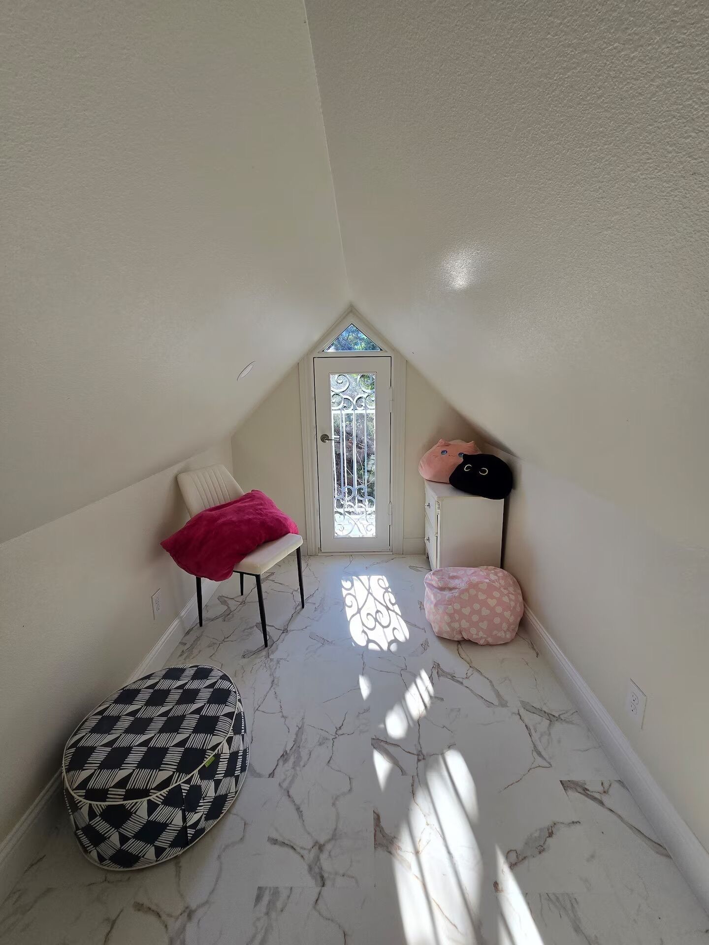 Um quarto com uma porta branca, uma cadeira com manta rosa, um banquinho estampado e uma almofada rosa.