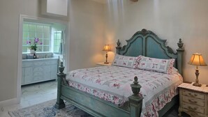 Une chambre à coucher avec un lit, deux tables de chevet avec des lampes, un lustre, une fenêtre donnant sur un beau paysage, et une salle de bain avec un meuble-lavabo et un miroir.