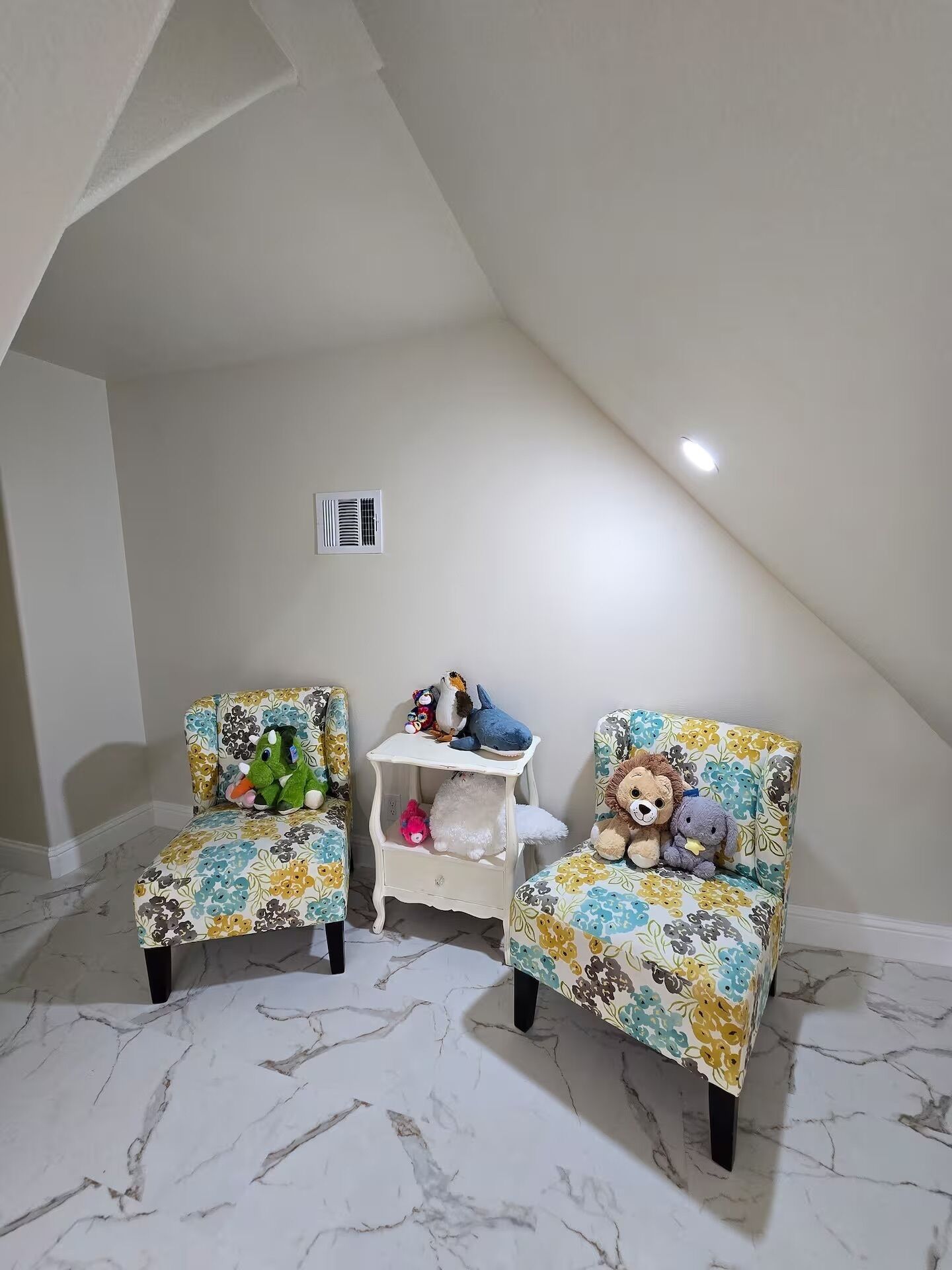 Een kamer met twee fauteuils met bloemenprint, een klein wit schap met knuffeldiertjes, en een wit plafond met een ventilatieopening.