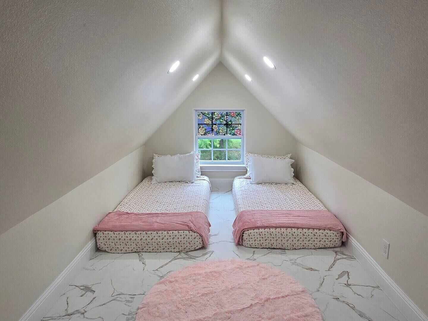Een kleine, driehoekige slaapkamer op zolder met twee bedden, een roze vloerkleed en een glas-in-loodraam.