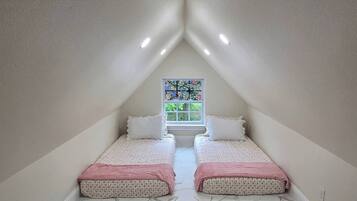 Een kleine, driehoekige slaapkamer op zolder met twee bedden, een roze vloerkleed en een glas-in-loodraam.