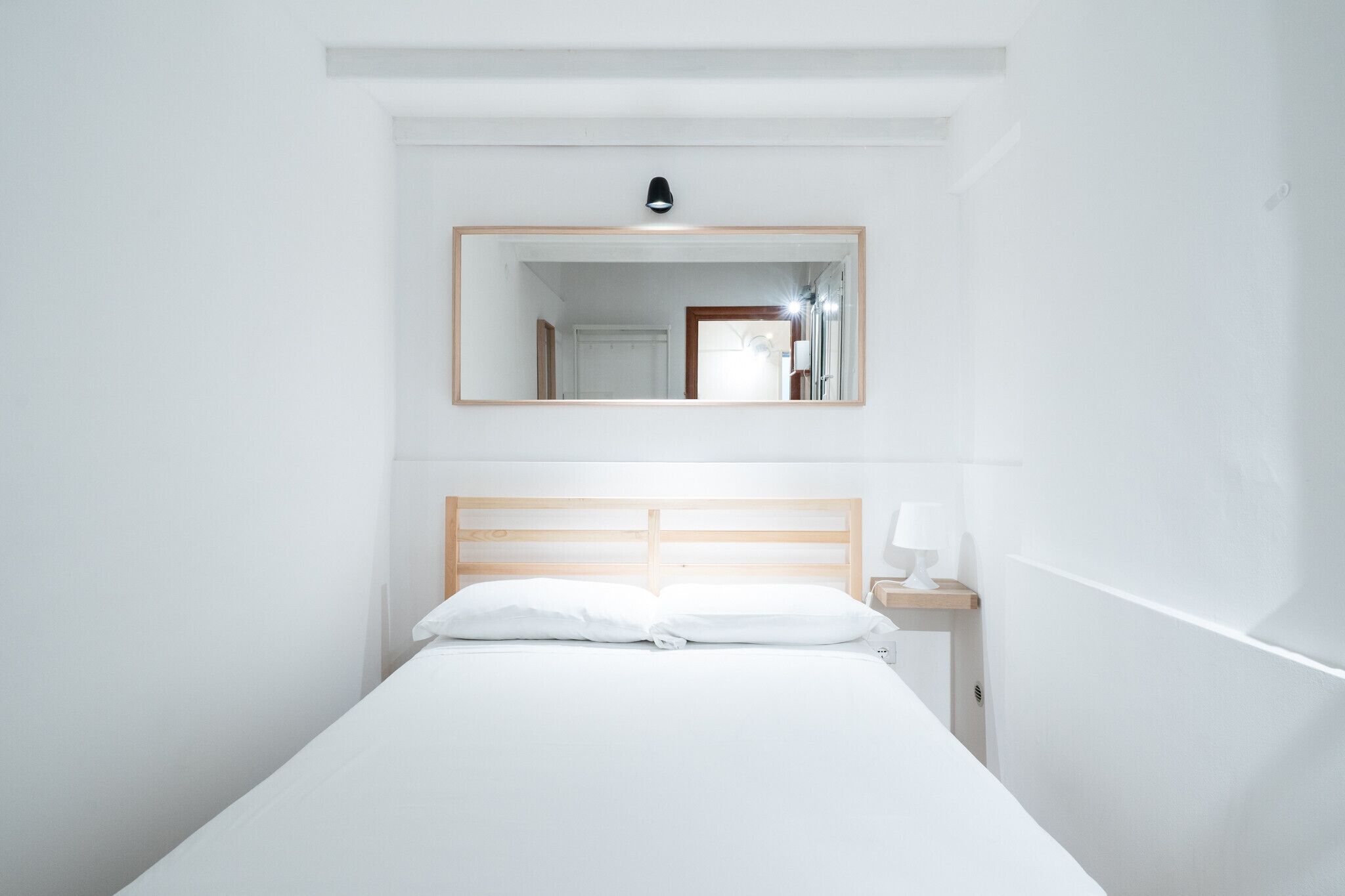 Habitación de hotel minimalista con una cama individual, una mesita de noche, un espejo y una lámpara fijada a la pared.