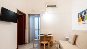 A small, well-lit room with a wooden dining table, four chairs, a beige sofa, a flat-screen TV mounted on the wall, and a painting on the wall. - Meta Apartment by Wonderful Italy (Bari)