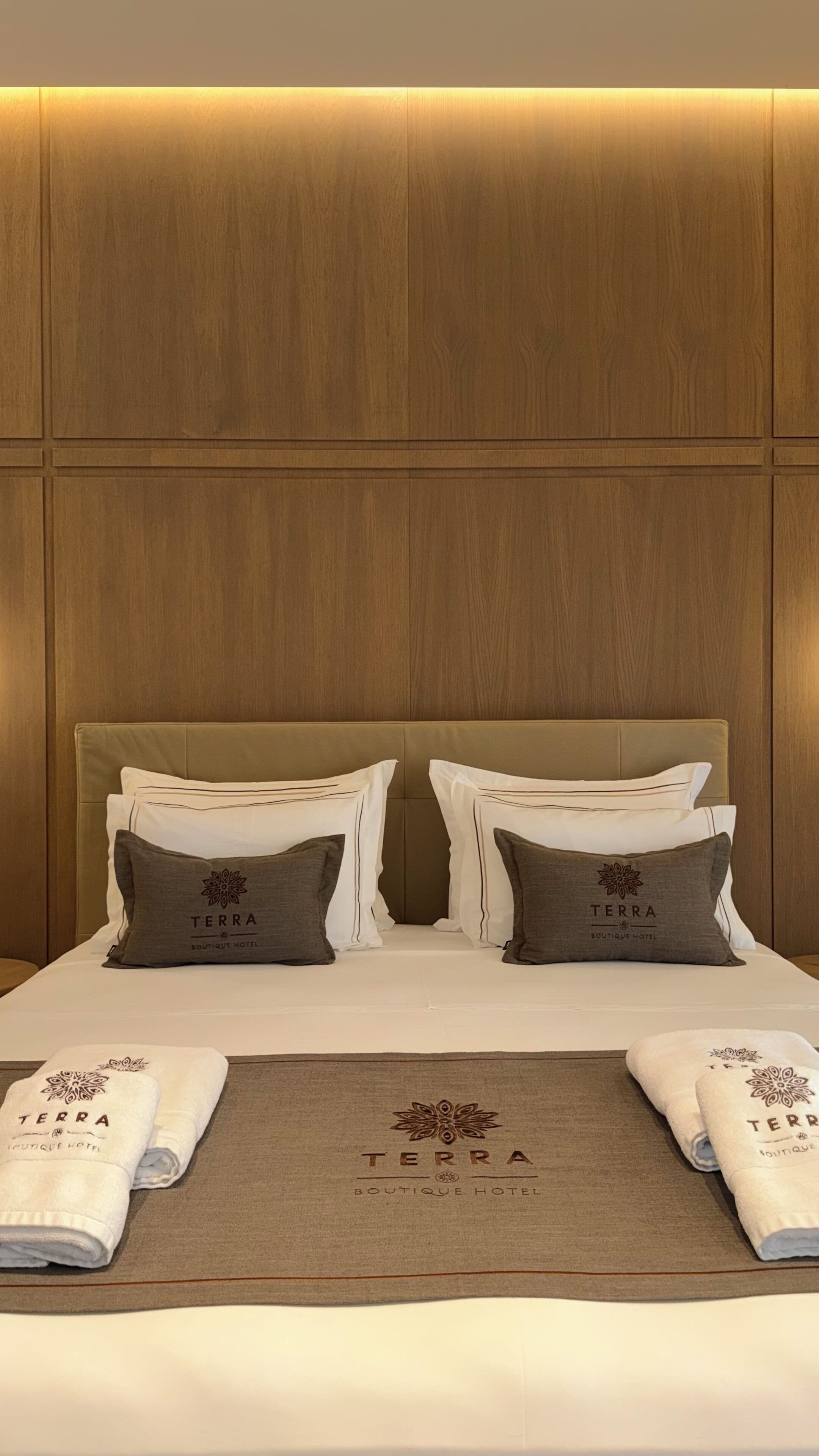 Habitación de hotel con cabecera de madera, dos almohadas con un logotipo, cama bien hecha con una toalla y un logotipo en la colcha.