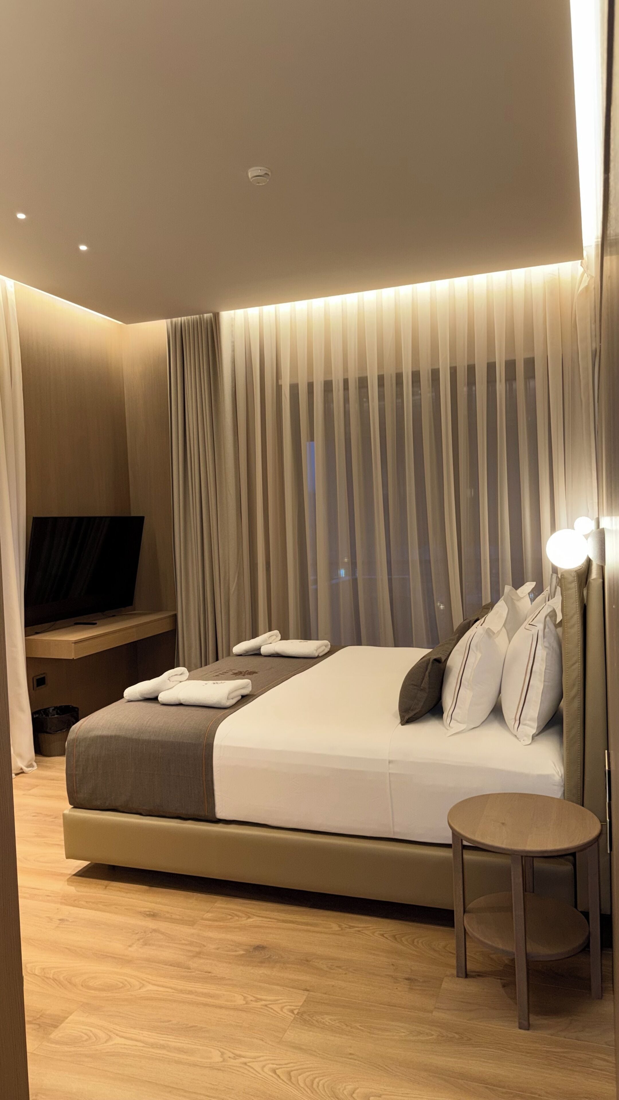 Habitació d'hotel moderna amb un llit gran, una taula de nit, una taula rodona petita, una televisió i cortines translúcides.