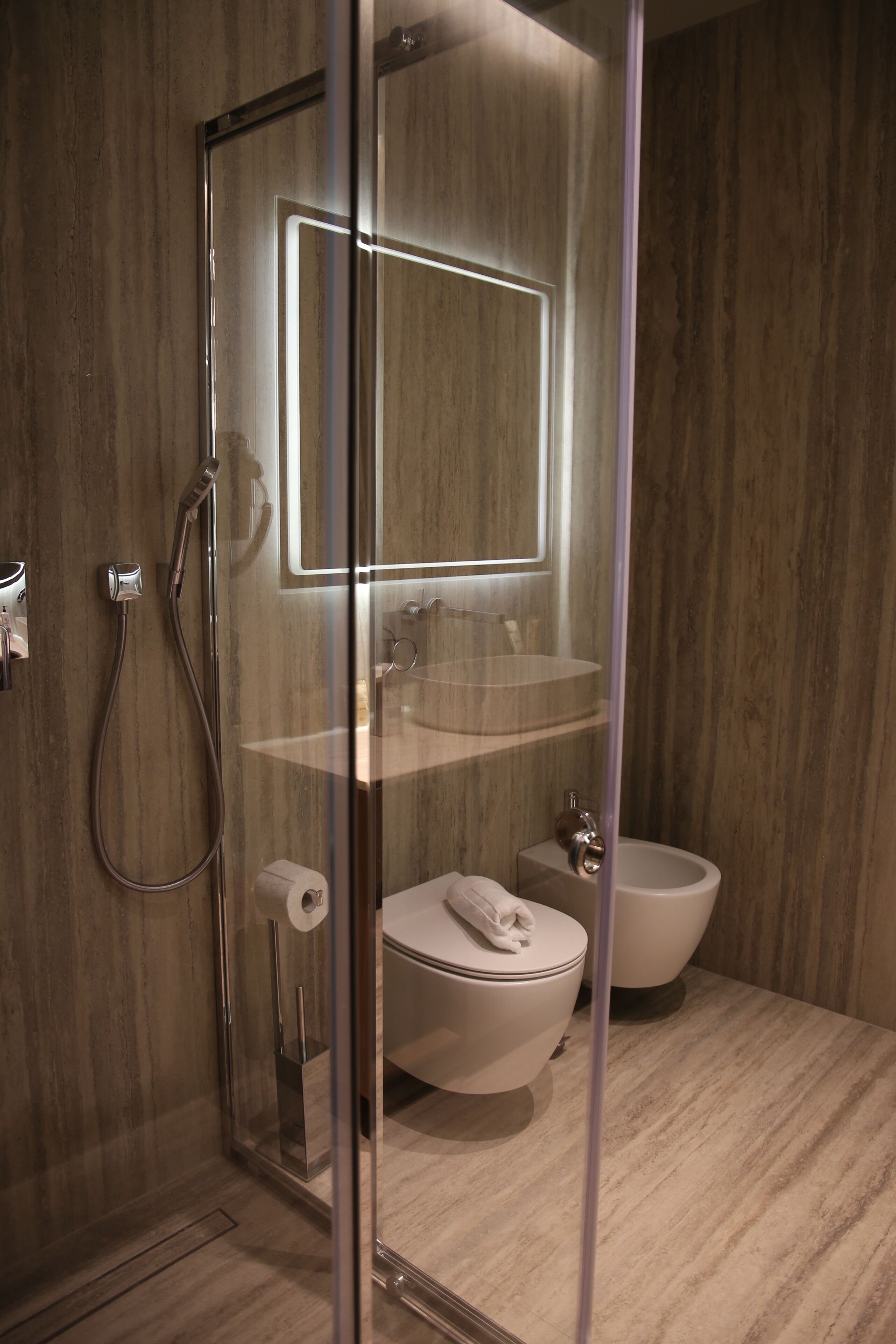 A modern bathroom with a glass-enclosed shower, a toilet, a bidet, and a mirror with integrated lighting.