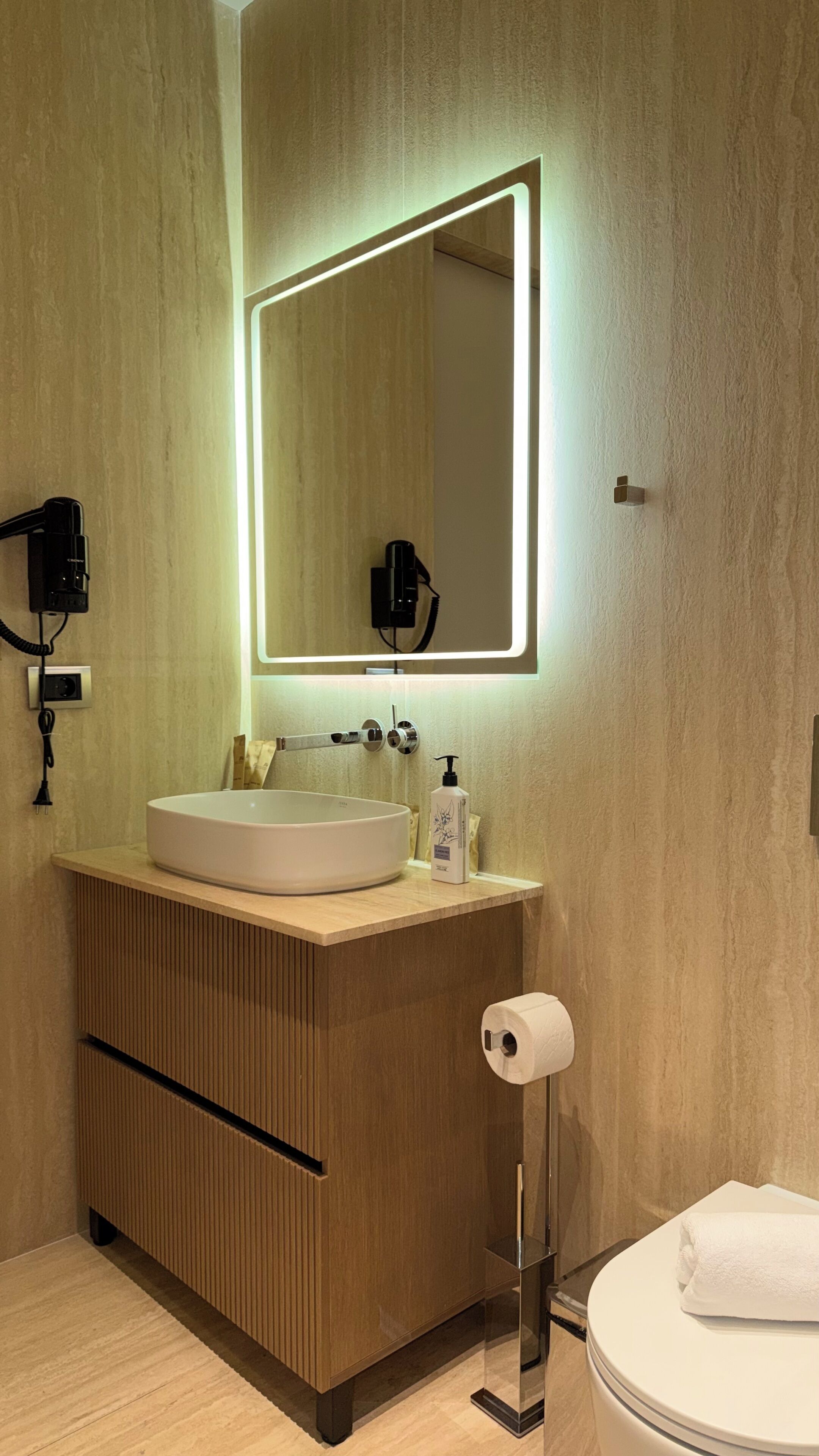 A modern bathroom with a wooden vanity, a large mirror with backlighting, a toilet, and a hand dryer.