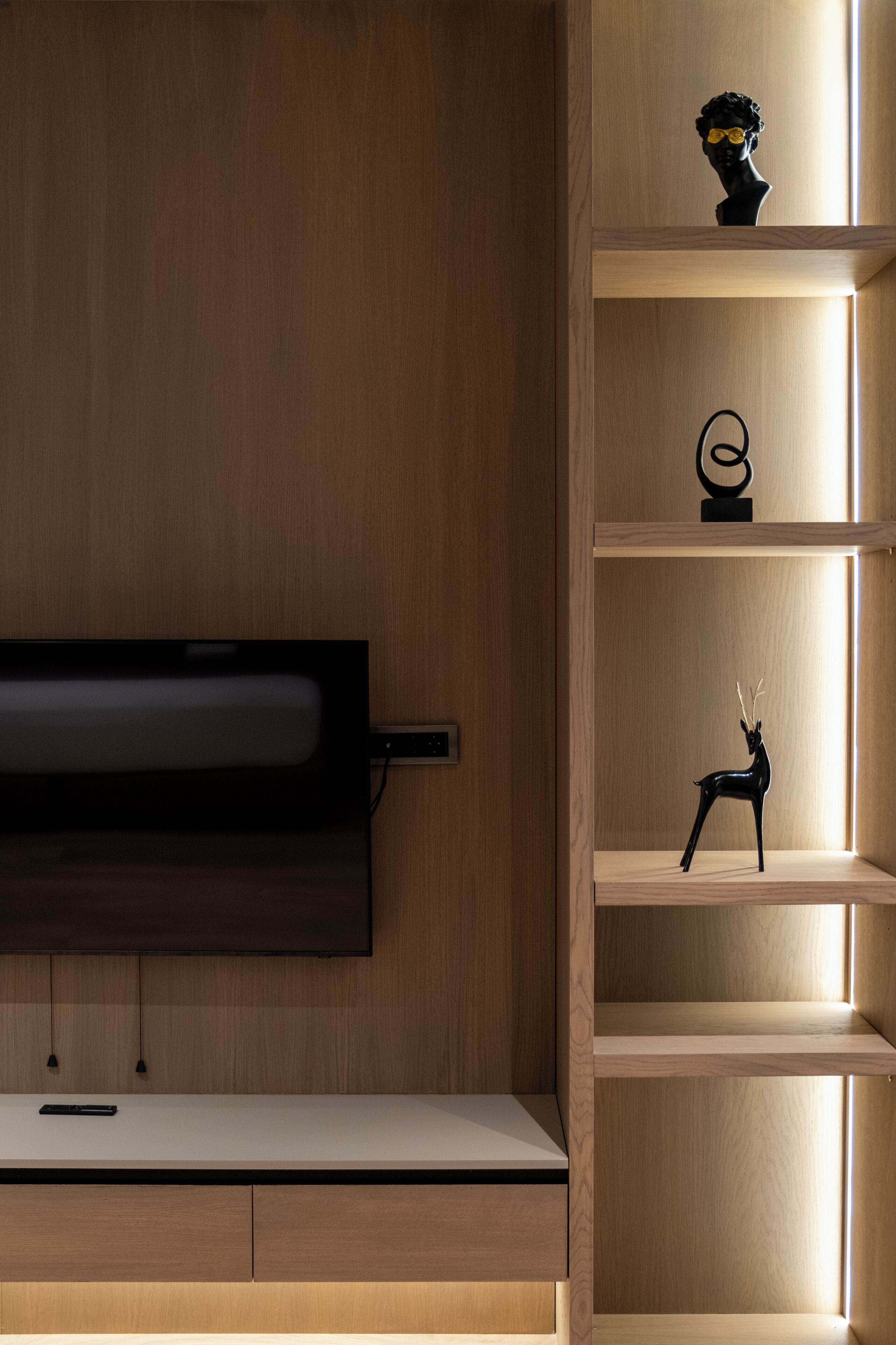 A modern room with a flat-screen TV mounted on a wooden wall, a wooden desk with drawers, and a shelf with decorative items.