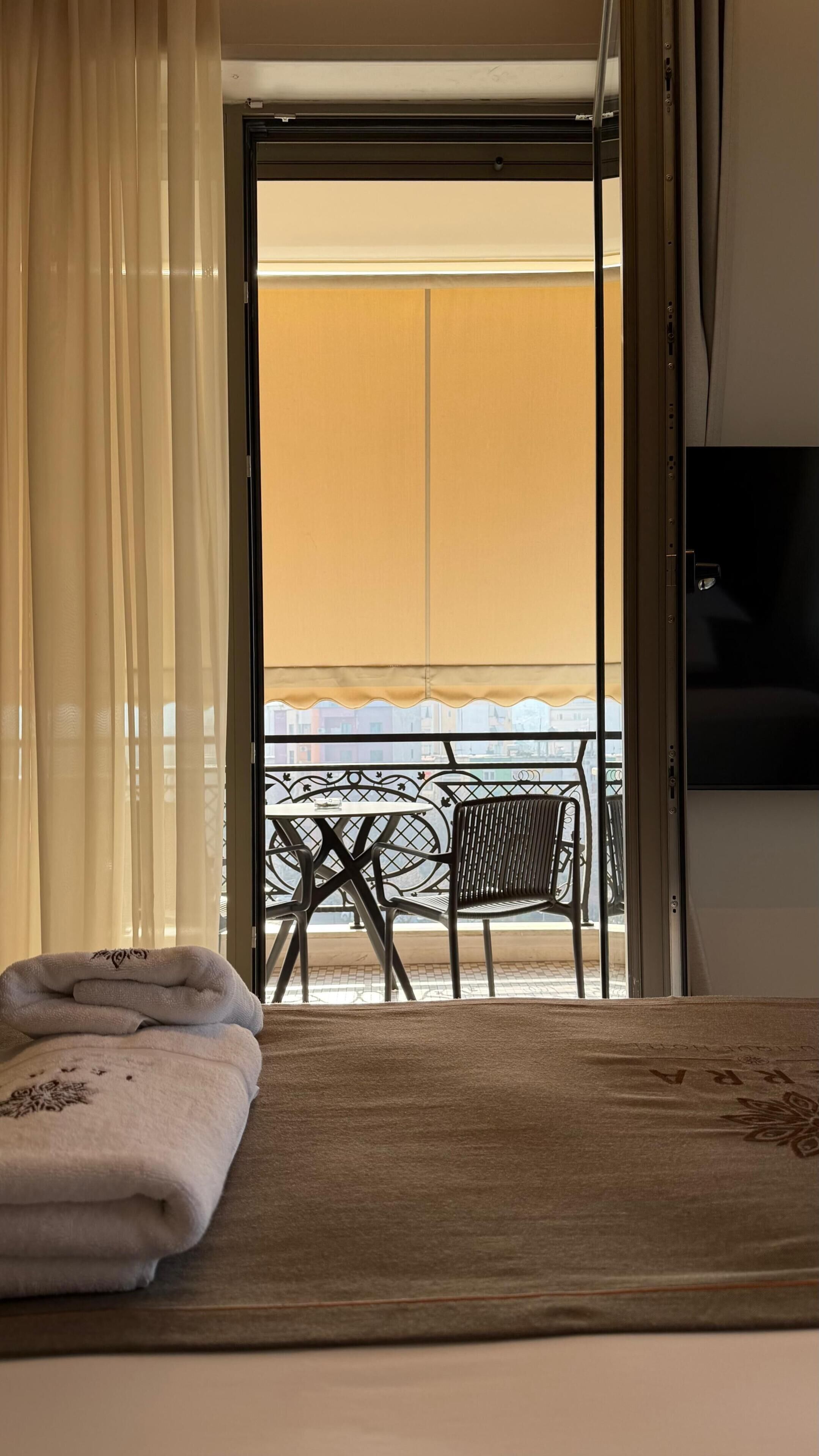 A hotel room with a bed, a TV, a balcony with a table and chairs, and a view of buildings.
