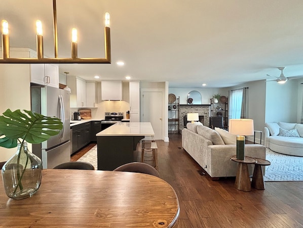 A modern kitchen with a central island, dining area with a round table, and a living room with a sofa and lamp.
