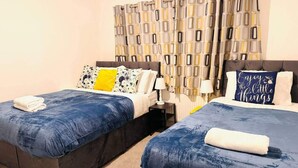 A hotel room with two beds, a nightstand, a lamp, and a patterned curtain. - Modern Easedale House - 3 Bed House in Hornchurch (Hornchurch)
