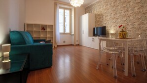 Een moderne woonkamer met een blauwgroene bank, een eettafel met transparante stoelen, een televisie en behang met een patroon.