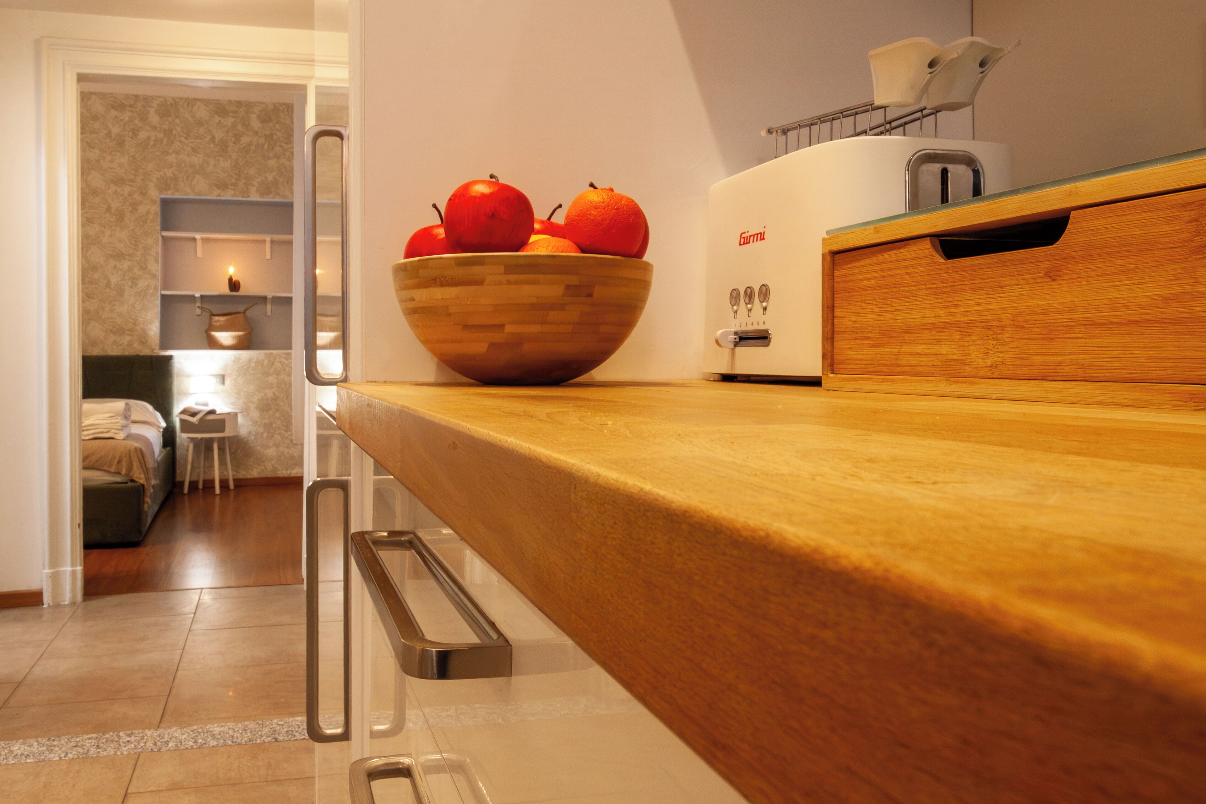 Une cuisine avec un plan de travail en bois, un saladier rempli de fruits, un grille-pain, et un aperçu d’une chambre à coucher avec un lit et une table de chevet.