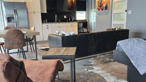 A modern kitchen with white cabinets, a black countertop, and a flat-screen TV. There is a dining area with a wooden table and brown chairs. A black sofa and ottoman are in the living area, and a rug covers the floor.