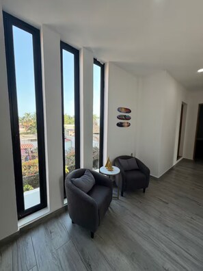 Una habitación amplia con ventanas grandes, un sofá gris, una mesita redonda y un sillón oscuro.