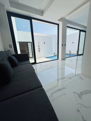 A modern interior with a sofa, large sliding glass doors leading to a pool area, and a clean, tiled floor.