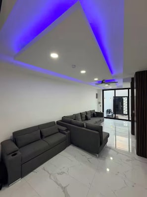 Uma sala de estar moderna com sofá modular, teto com iluminação embutida e uma grande porta de vidro que dá para uma área externa.