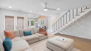 Un amplio salón con un sofá seccional color beige, un pequeño reposapiés y una escalera con detalles de madera.