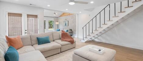 Un salon spacieux avec un canapé d’angle beige, un petit pouf et un escalier avec des finitions en bois.