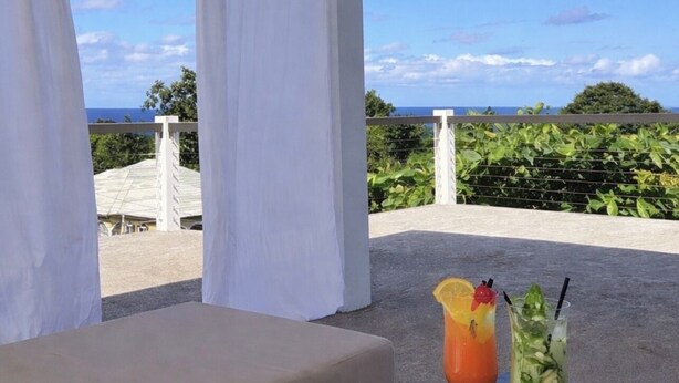 A balcony with a view of the ocean, a lounge chair, and two drinks on a table. - Purevibe 2 BR/2 BA, Ocean View Villa, Private Rooftop, Near Port Antonio (Snow District)