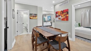 A dining area with a wooden table and chairs, a painting on the wall, and a view into a living room with a bed and a couch.