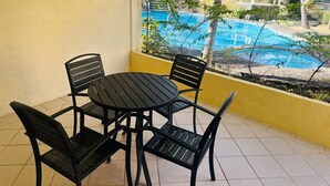 A patio with a round table and four chairs, overlooking a pool area with palm trees and a red-roofed building.