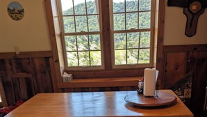 Una mesa de madera para comer con bancos, una ventana que da a un bosque y un reloj colgado en la pared.