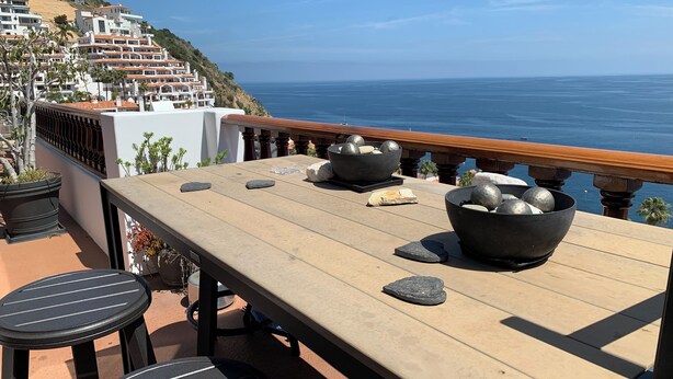 Una terraza con vista al océano, que incluye una mesa con cuencos y piedras, y una barandilla que da al pie de una colina con edificios.