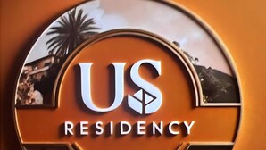 Un logo circulaire avec le texte « US RESIDENCY » et une vue partielle d’un palmier et d’un paysage de plage.