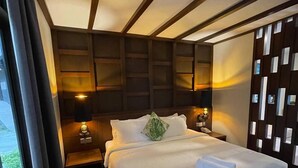 A hotel room with a large bed, wooden headboard, bedside lamps, and a view of greenery outside.