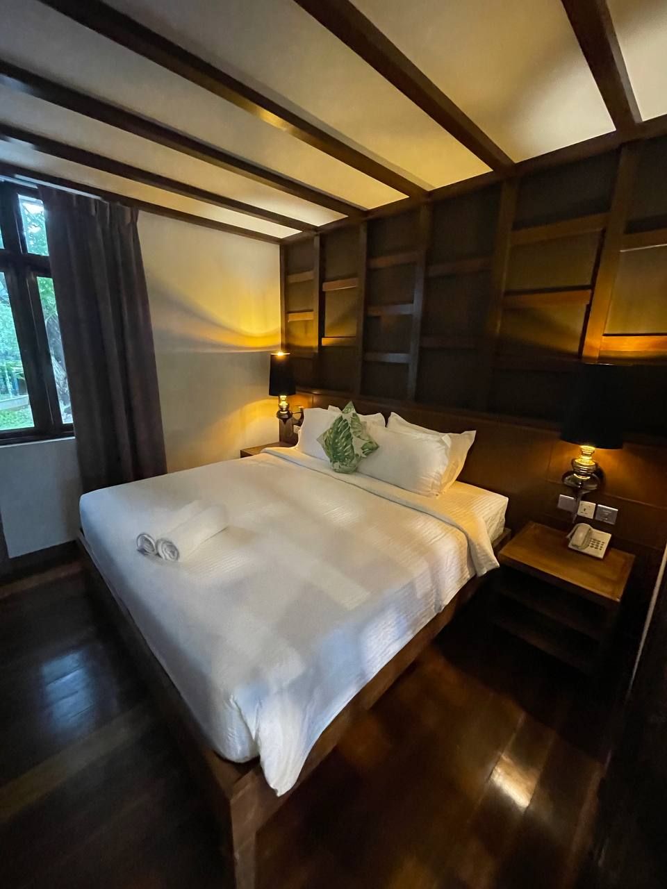 A neatly made bed with a white comforter and pillows, a bedside table with a lamp, and a window with curtains.