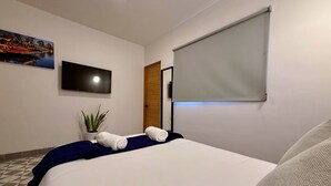 Et hotelværelse med en seng, et vægmonteret fladskærms-TV, en potteplante og et vindue med rullegardin.