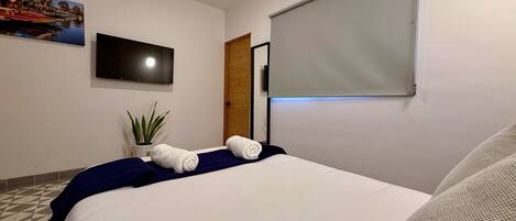 Habitación de hotel con una cama, un televisor de pantalla plana en la pared, una planta en maceta y una ventana con persiana.