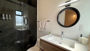 Et moderne badeværelse med et glasafskærmet brusebad, et hvidt toilet, en trækommode med en hvid vask, et rundt spejl og en håndklædestang.