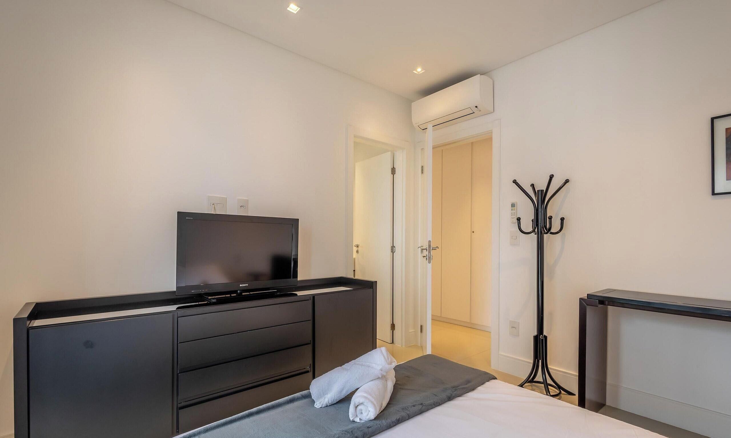 Habitación de hotel con televisor de pantalla plana sobre un tocador, cama con manta gris, perchero y puerta que da a otra habitación.
