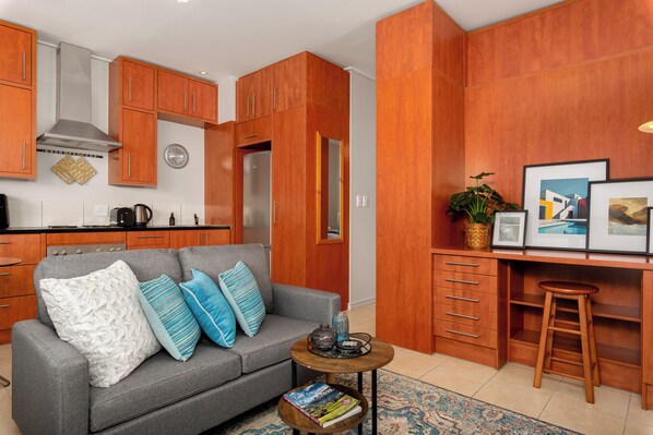 A modern kitchen with wooden cabinets and stainless steel appliances, a grey sofa with patterned cushions, and a small wooden table with a book and a plant.
