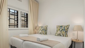 Un lit bien fait, avec du linge de lit blanc et un plaid beige, une table de chevet avec une lampe, et une fenêtre avec des stores et des rideaux.