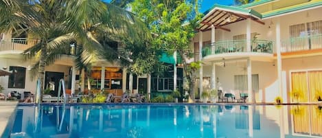 Una piscina con superficie de agua azul cristalina, rodeada de palmeras y un edificio de dos pisos con balcones.