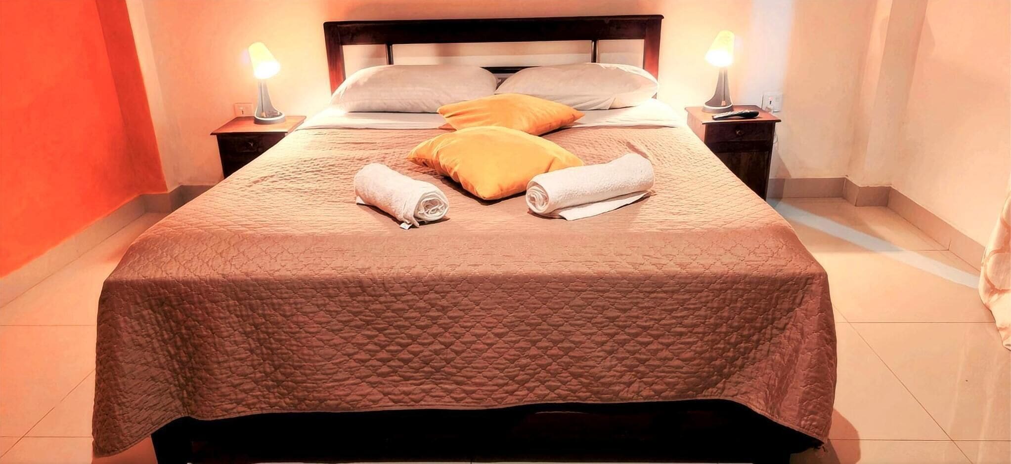 Ein ordentlich bezogenes Bett mit einer gesteppten Decke, zwei Kissen und gerollten Handtüchern. Auf dem Nachttisch stehen zwei Nachttischlampen und eine Fernbedienung.