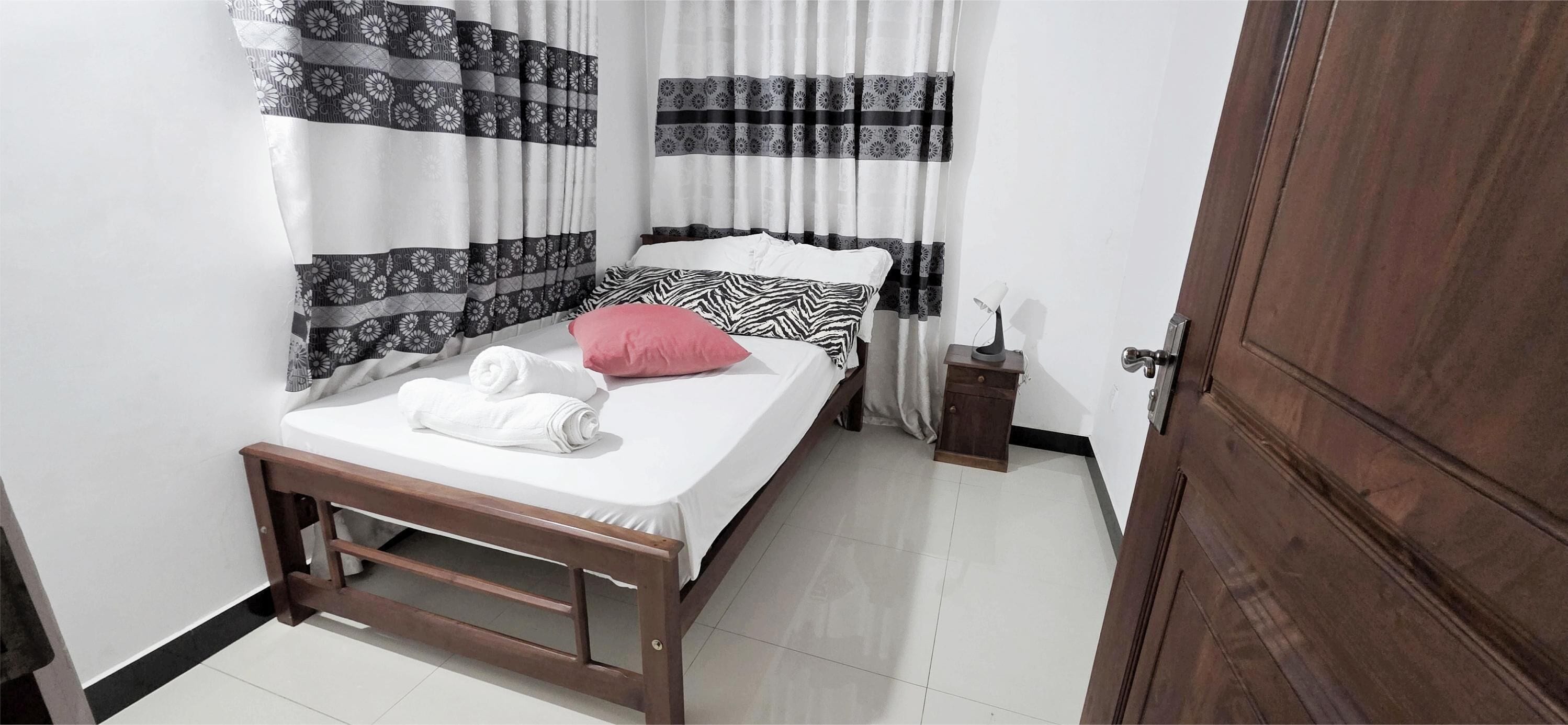 A neatly made bed with white and pink bedding, a bedside table with a lamp, and patterned curtains.