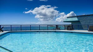 A clear blue pool with a view of the ocean, a fenced deck area with chairs, and a building with a green roof.
