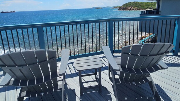 A deck with Adirondack chairs and a table overlooking a beach with a ship in the distance.