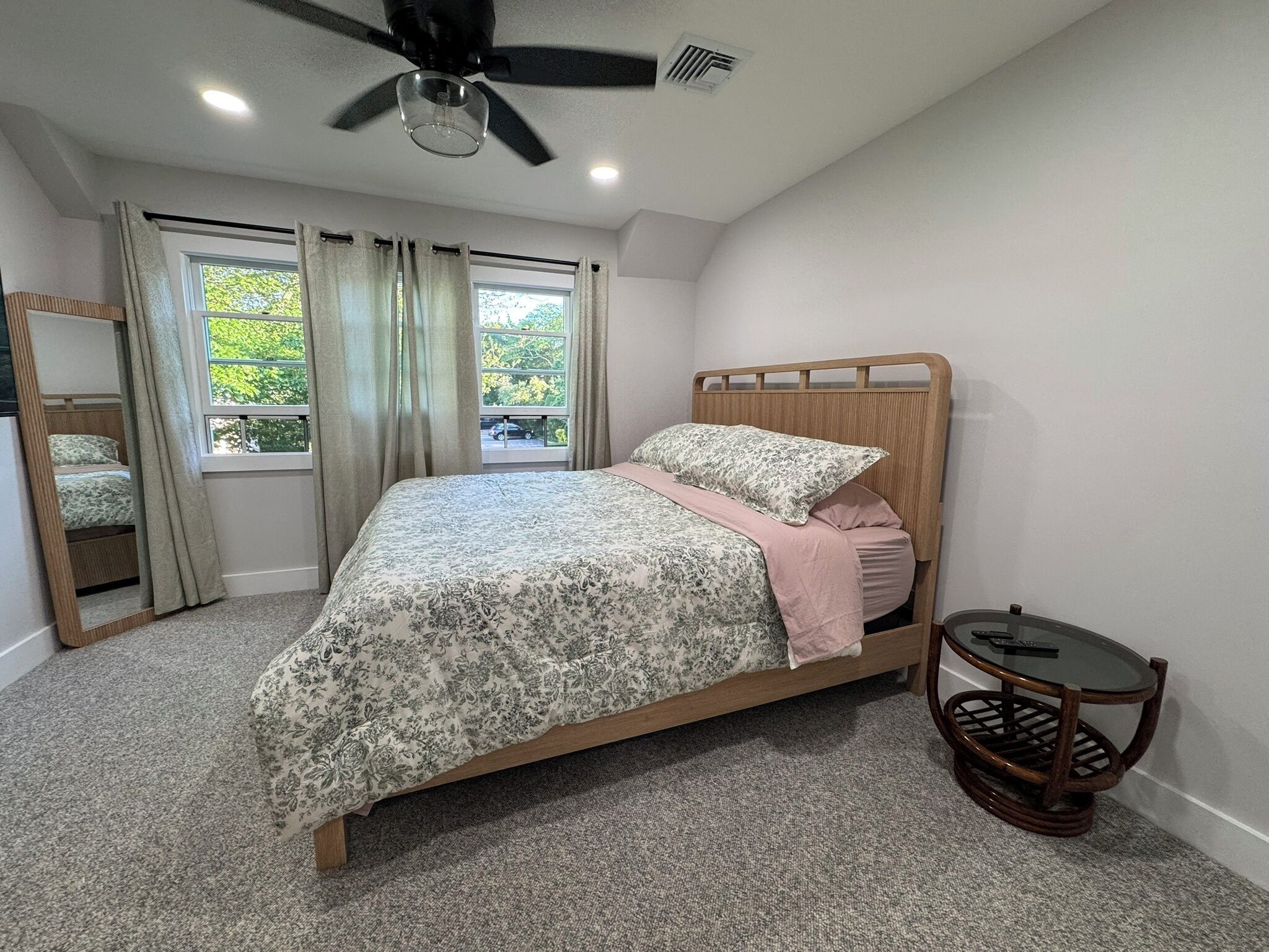 A bedroom with a bed, a ceiling fan, a window with curtains, a small table, and a mirror.