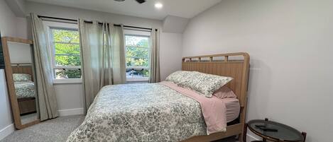 A bedroom with a bed, a ceiling fan, a window with curtains, a small table, and a mirror.