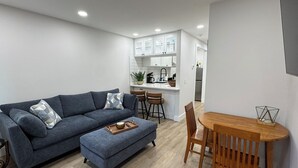 A modern living room with a grey sofa, wooden dining table, and a kitchen in the background. - Heart of the Hamptons, Waterfront with Pool!  1 Bed/1 Bath Townhome (Hampton Bays)