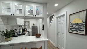 A modern kitchen with white cabinetry, a central island with two bar stools, a refrigerator, and a sink. - Heart of the Hamptons, Waterfront with Pool!  1 Bed/1 Bath Townhome (Hampton Bays)